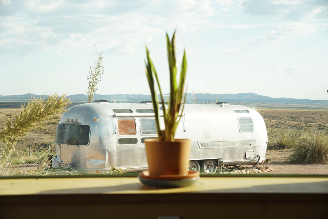 Vintage Airstream trailer parked in a serene desert landscape, seen through a window with a potted plant in the foreground. Represents self-care, embracing limitations, and the peaceful journey of living with chronic illness.